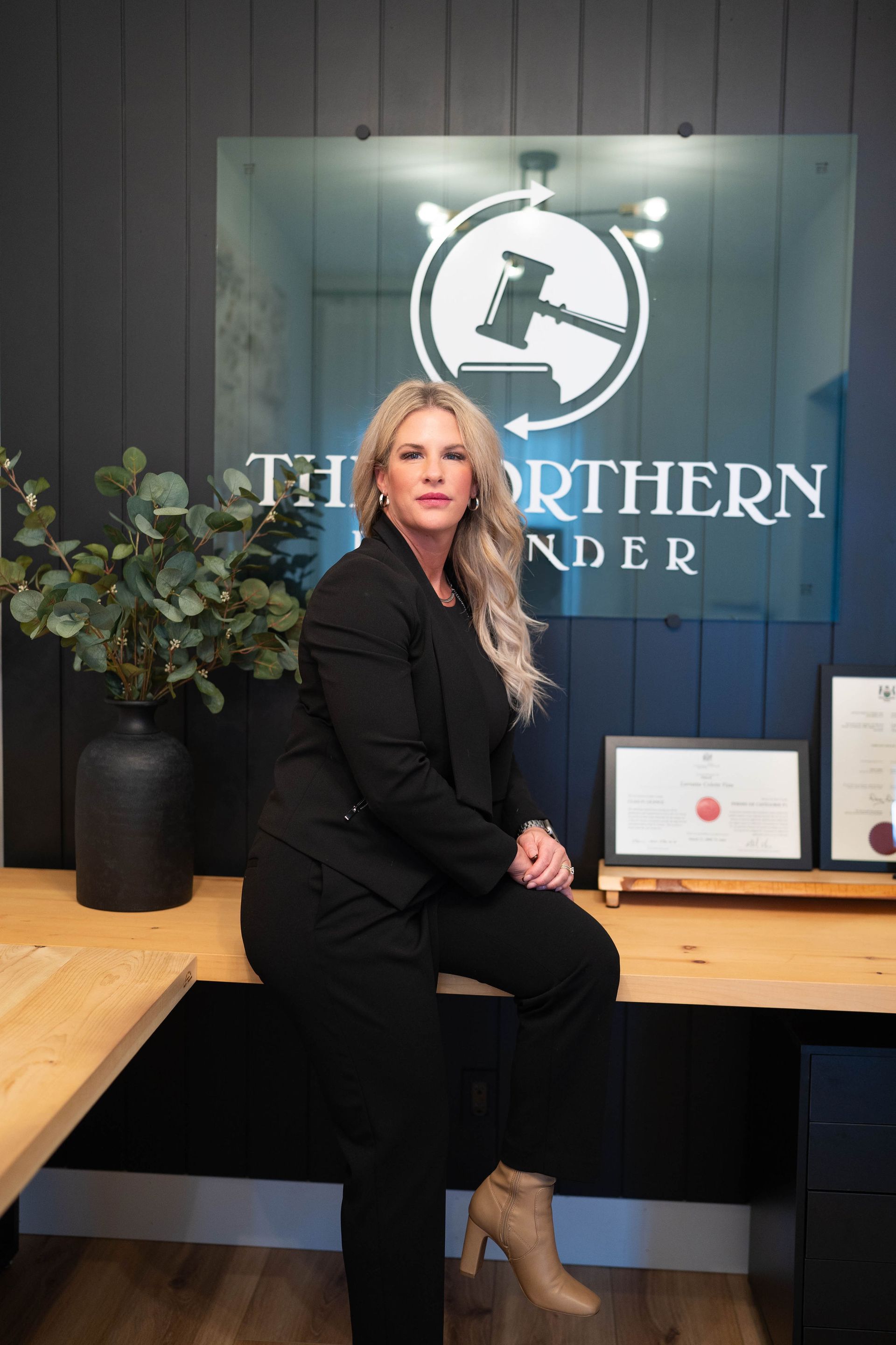 Woman in black suit posing at a desk with framed awards and a Northern Builder sign behind her.