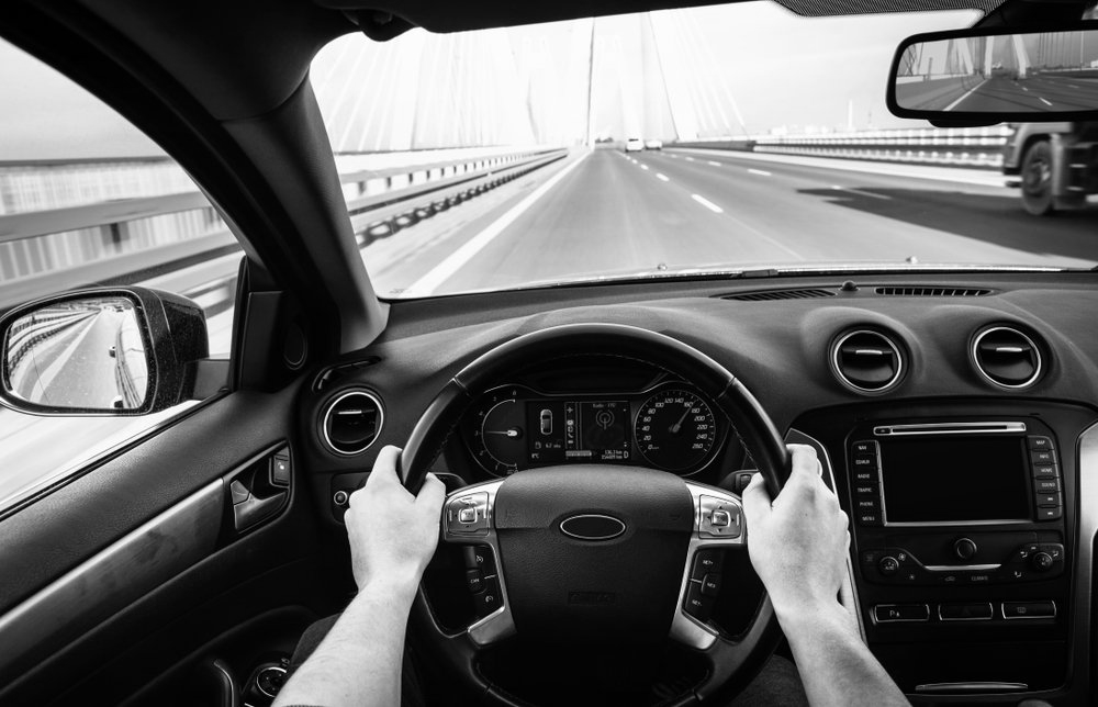 Driver's view from a car on a bridge, hands on the wheel, road stretching ahead in black and white.