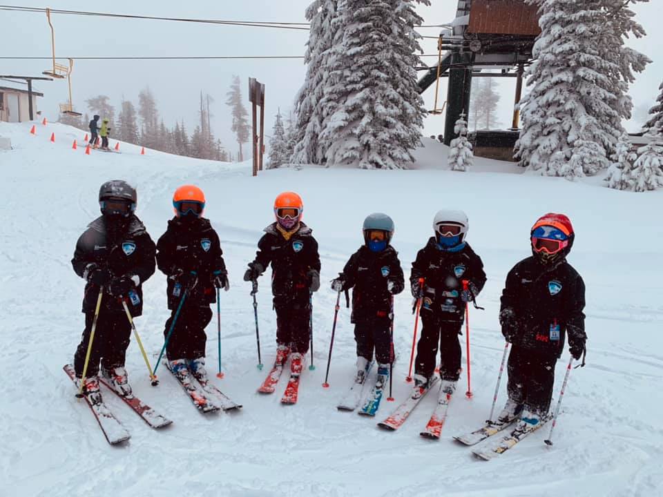 Six skiers in helmets and goggles lined up on a snowy ski slope near a chairlift.