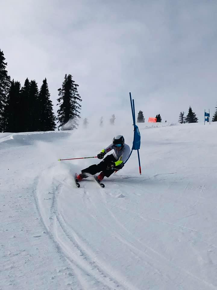 Skier carving a sharp turn on a snowy slope beside a blue gate, with trees and cloudy sky behind.
