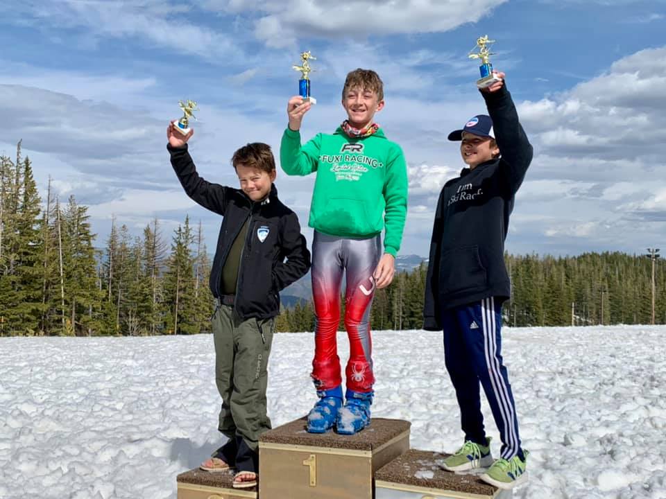 Three skiers on a snowy podium holding trophies in a winter landscape