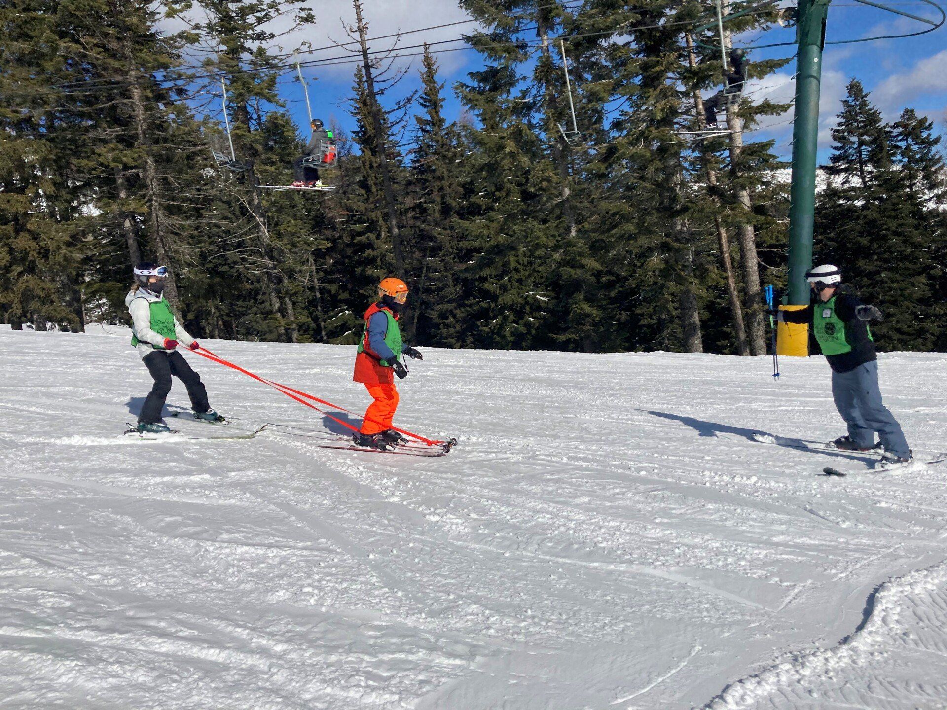 Three skiers in colorful gear descend a snowy slope with pine trees and a ski lift in the background.