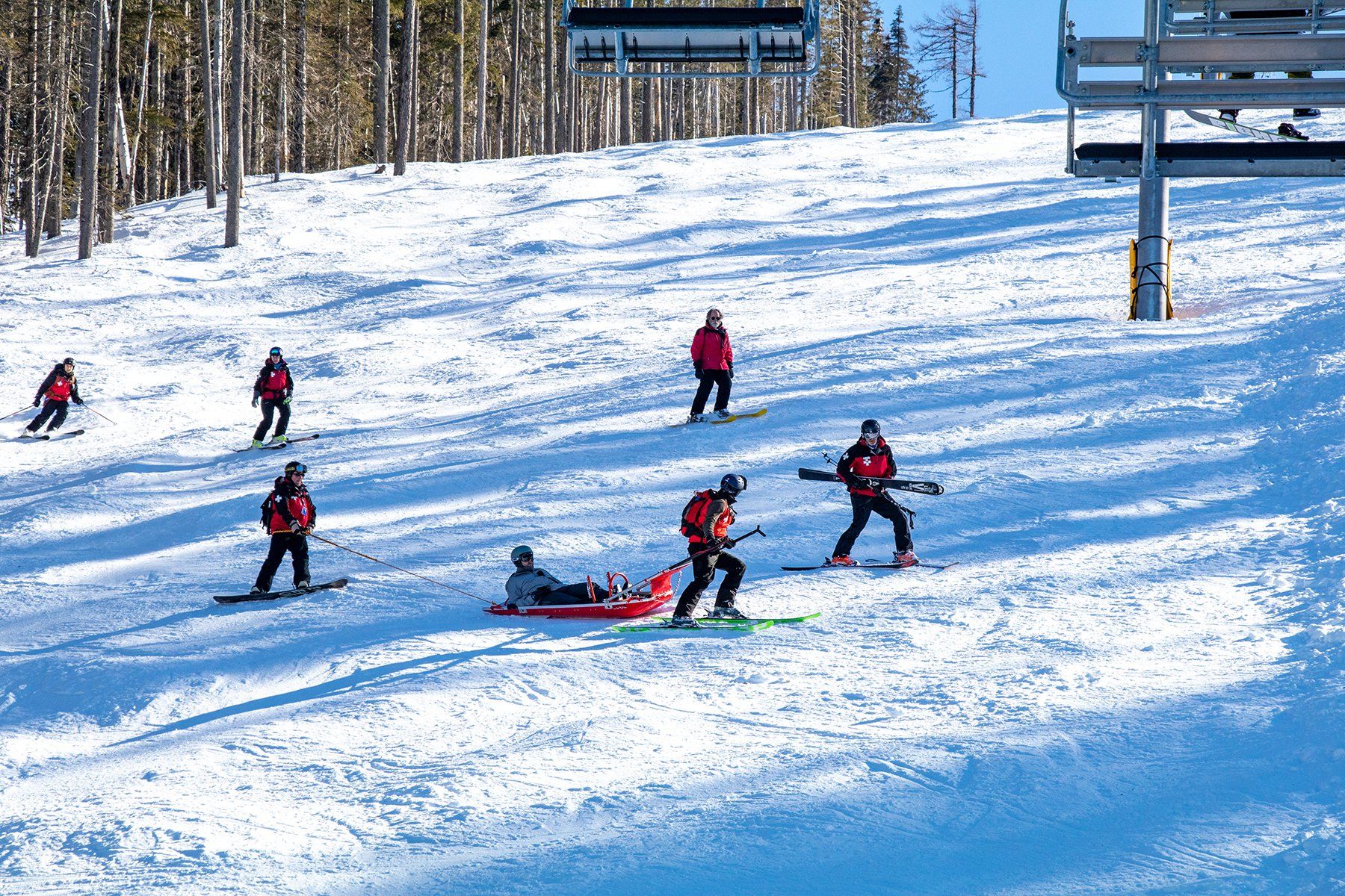 Skiers in red jackets descend a snowy slope, with one person helping another near the bottom.