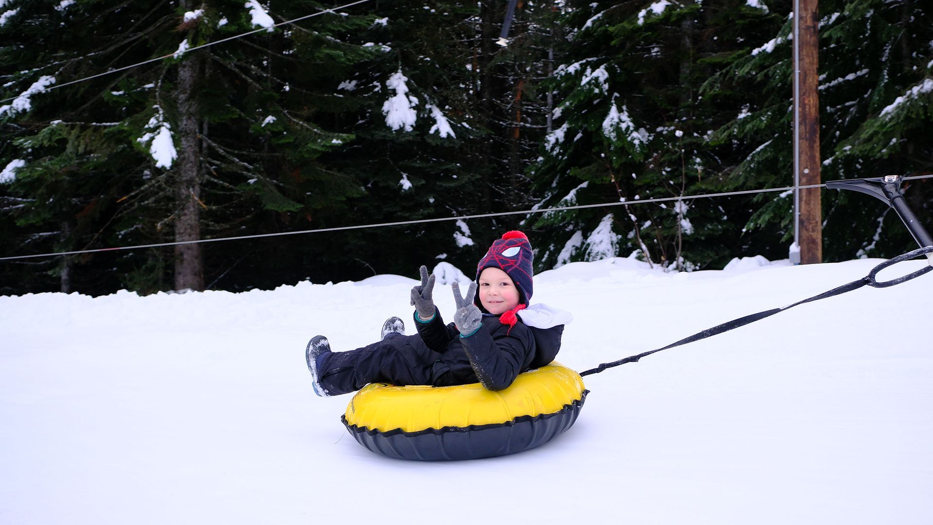 Child in a red hat riding a yellow snow tube down a snowy hill with trees in the background