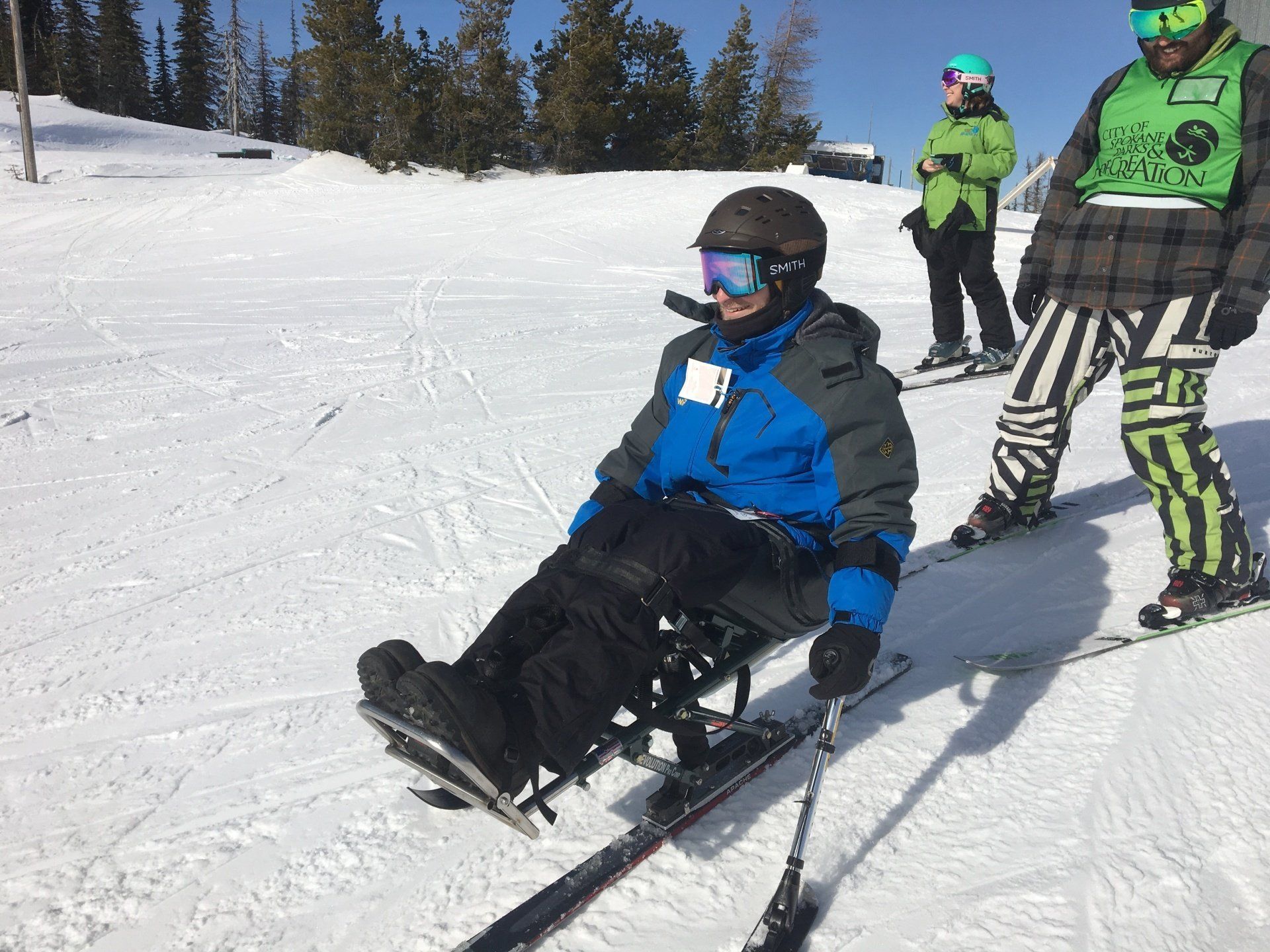 Skiers in green helmets watch a person in a blue jacket sled downhill on a snowy slope.