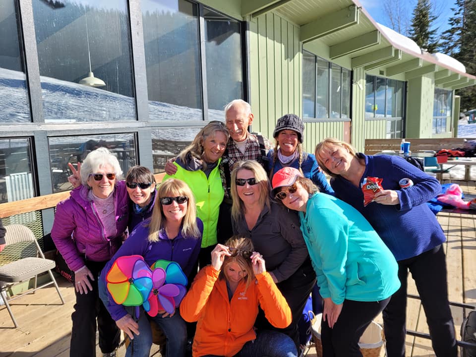 Group of people posing and smiling outdoors by a glass-walled building on a sunny deck.