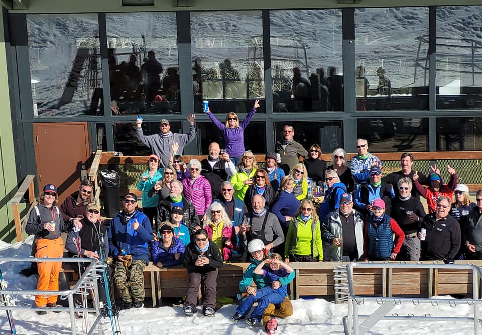 Group of people in colorful ski gear posing on snow outside a glass-front lodge, smiling and cheering