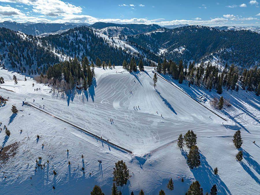 Snow-covered ski slope with trees and mountain ridges under a partly cloudy blue sky