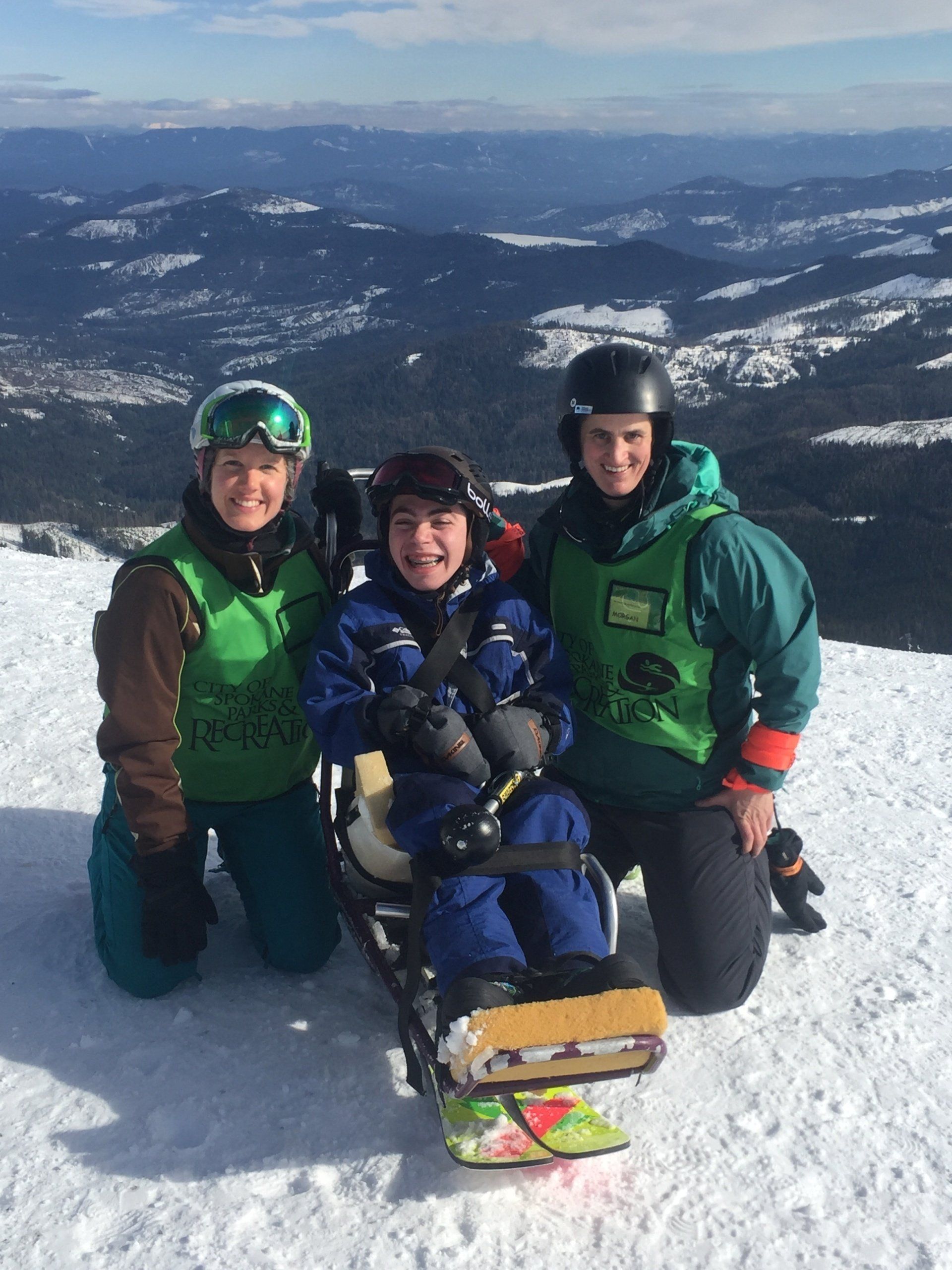 Three skiers kneel on a snowy mountain summit with skis and distant peaks behind them.