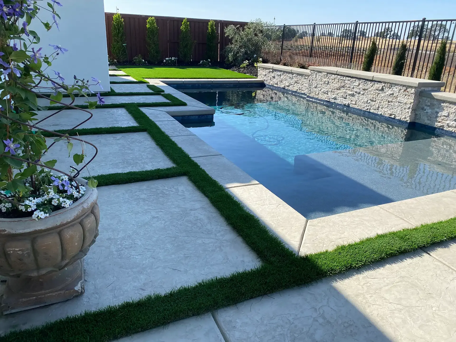 A rectangular pool with light blue water and stone borders, surrounded by concrete pavers and artificial turf.