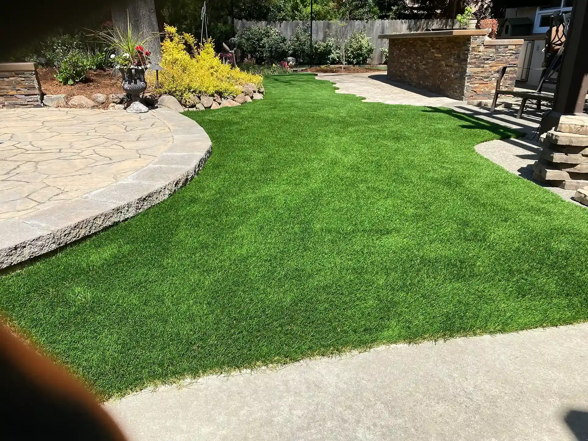 Green turf lawn bordered by concrete and a stone patio, with a garden bed and a stone bar in the background.