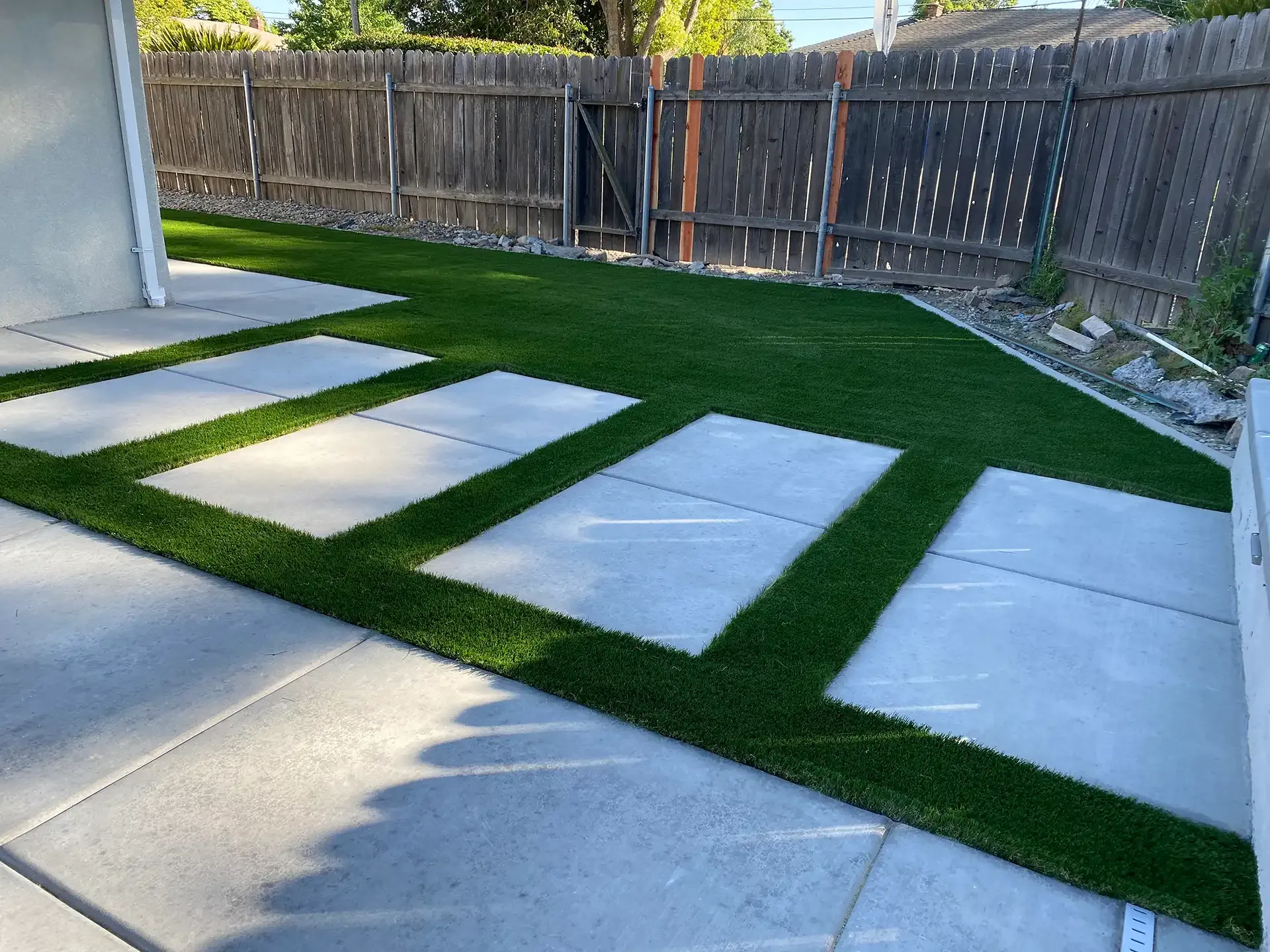 Backyard with square concrete stepping stones and green turf, bordered by a wooden fence.