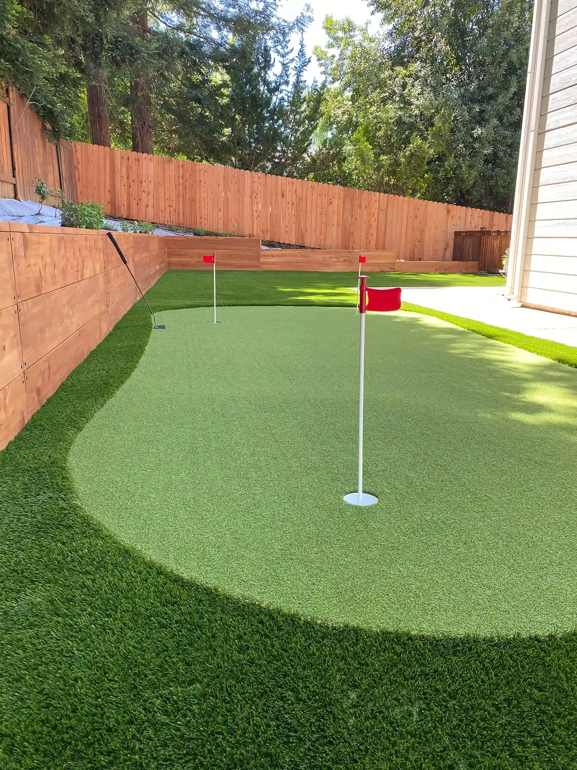 Putting green with two holes and red flags. Artificial turf with wooden retaining wall and fence.