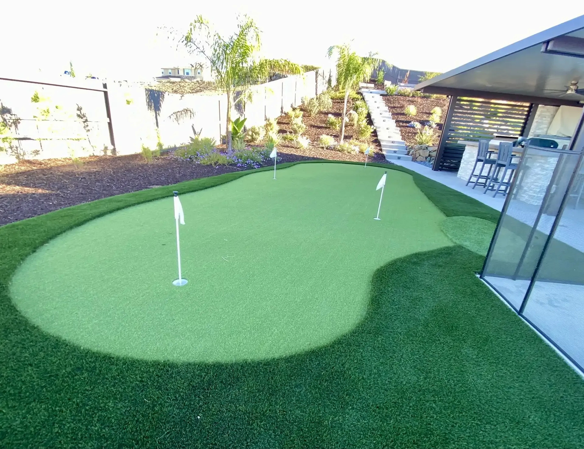 A backyard putting green with three flags, surrounded by artificial turf and landscaping near a patio.