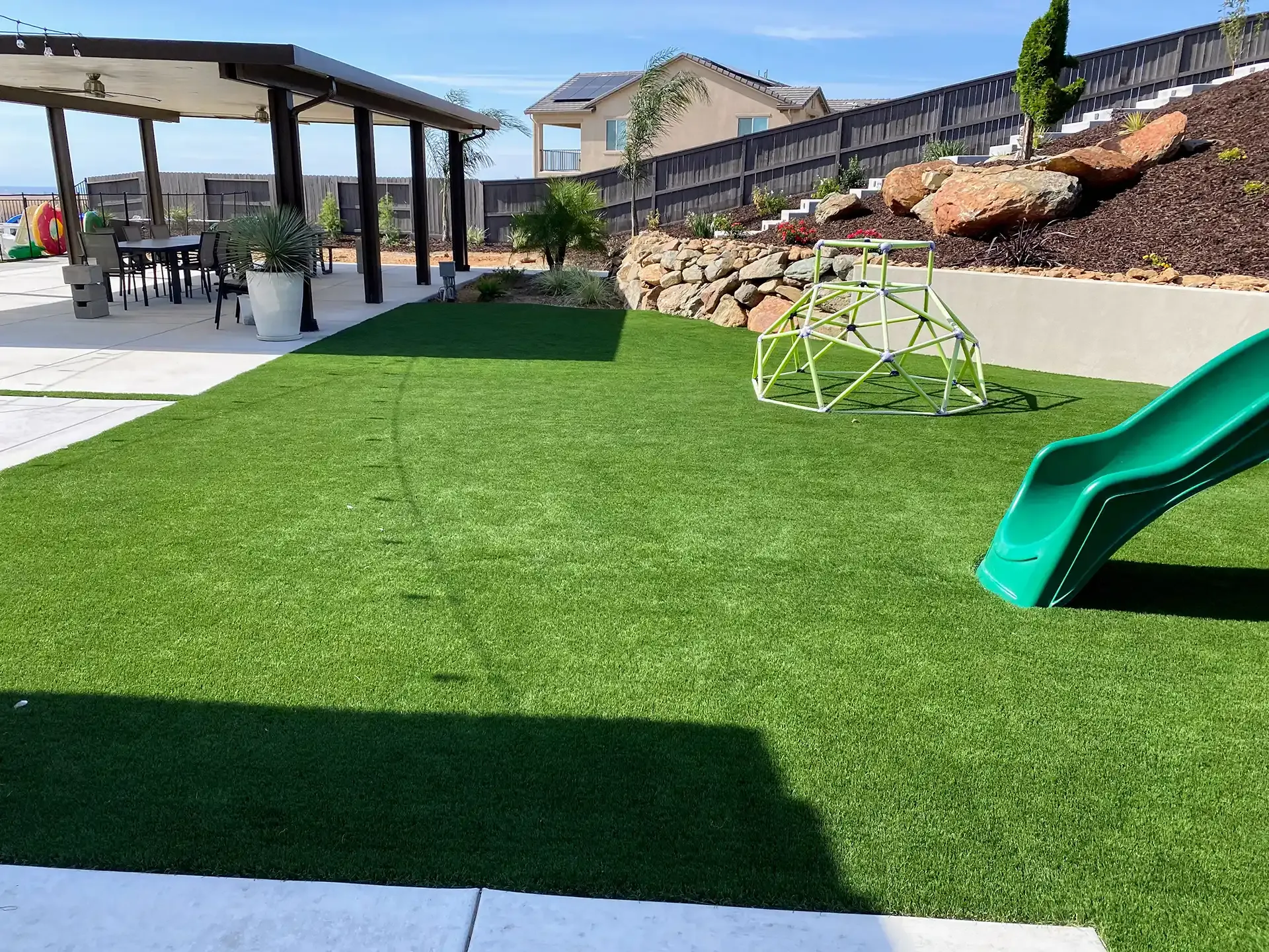 Backyard with green turf, play equipment, patio with table, and a house in the background.