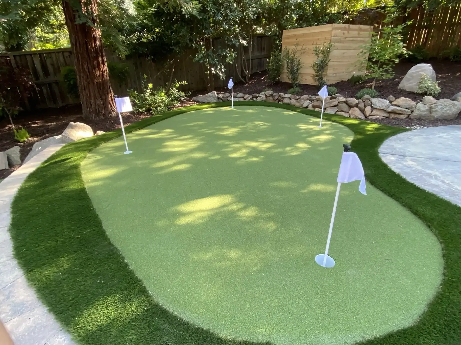 Artificial grass putting green with four holes, flags, and a sand trap in a backyard setting.