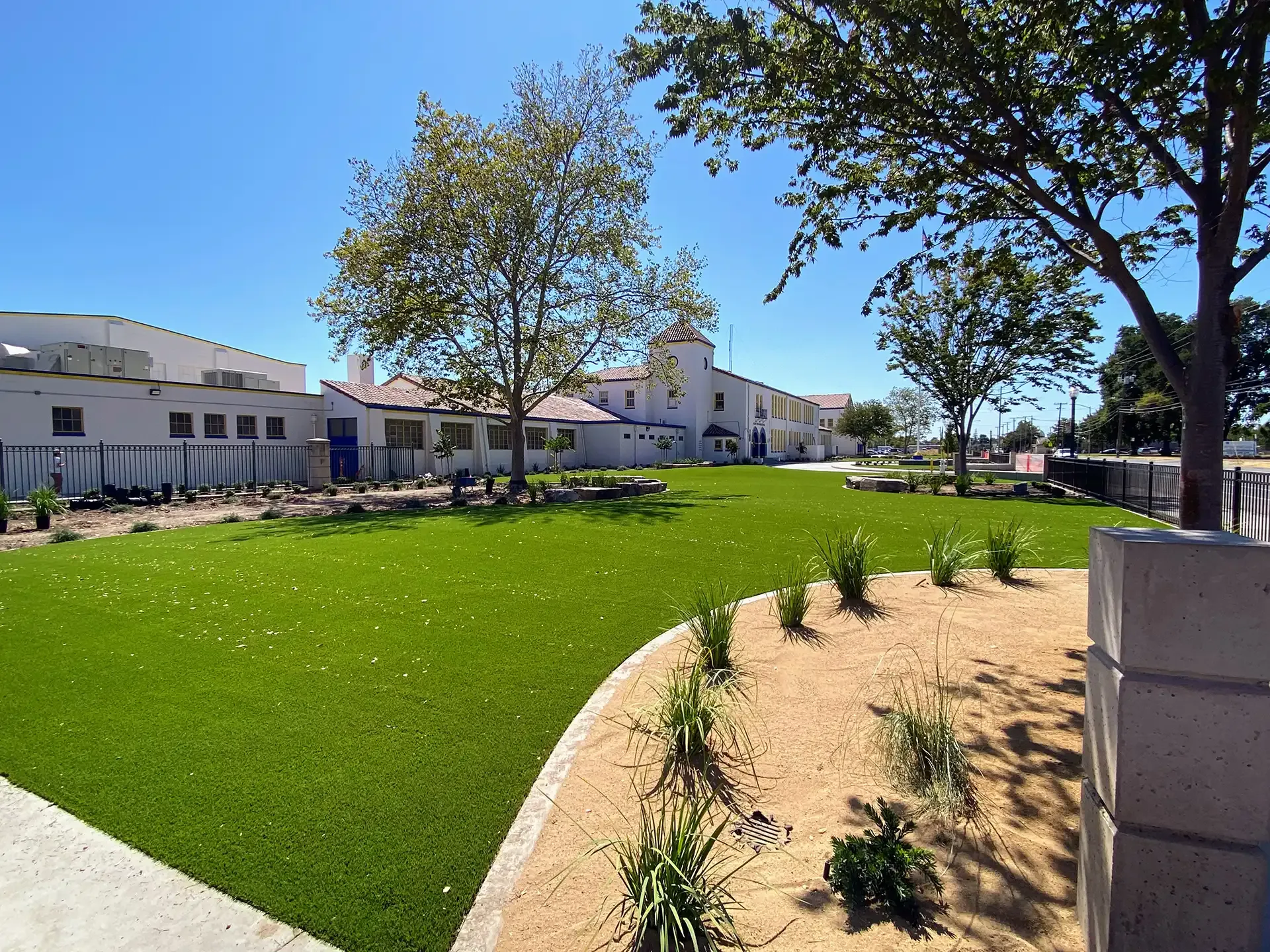 Green lawn and planted bed in front of a white building with trees under a blue sky.