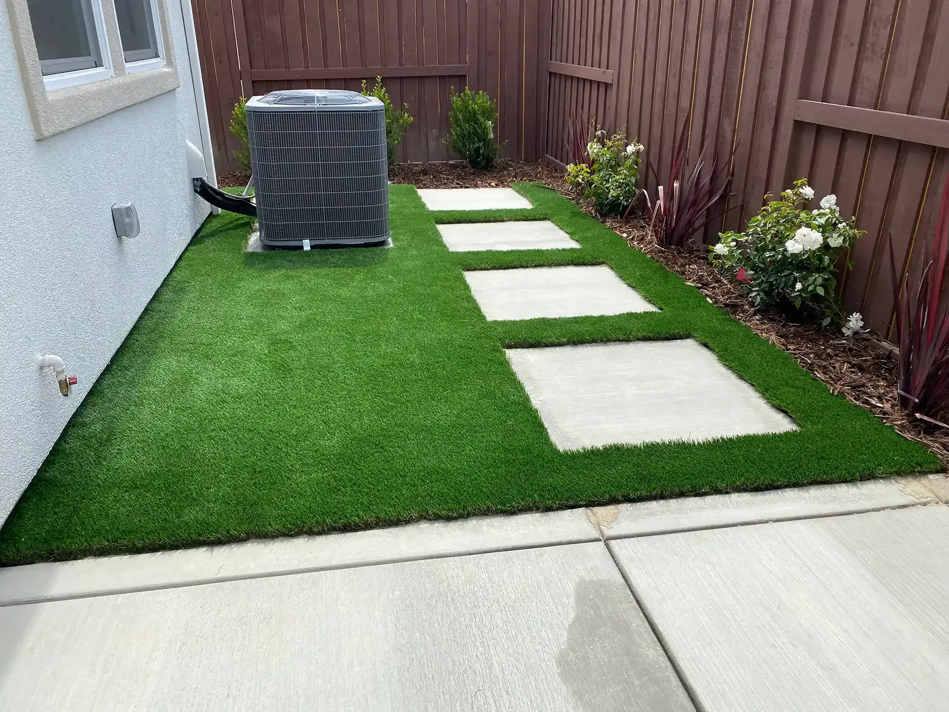 Artificial turf with stone pathway and air conditioning unit beside a house.