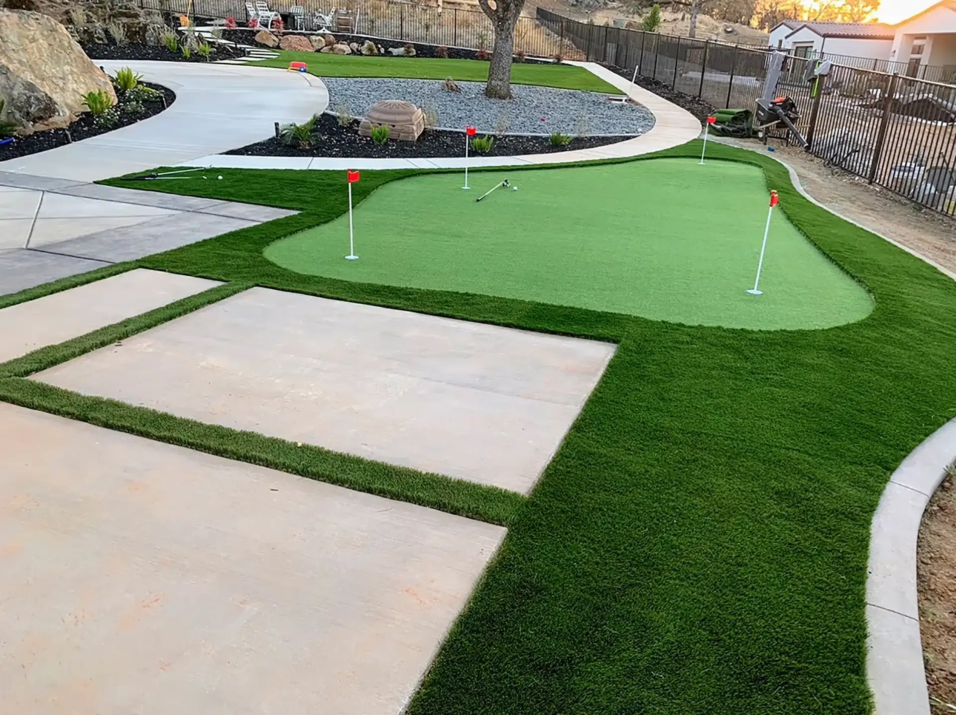 Green artificial turf and putting green in a landscaped backyard with concrete walkways.
