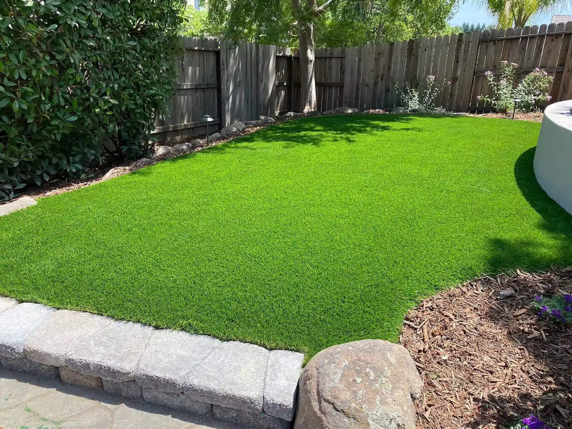 Green artificial turf in a backyard with a wooden fence and garden bed.