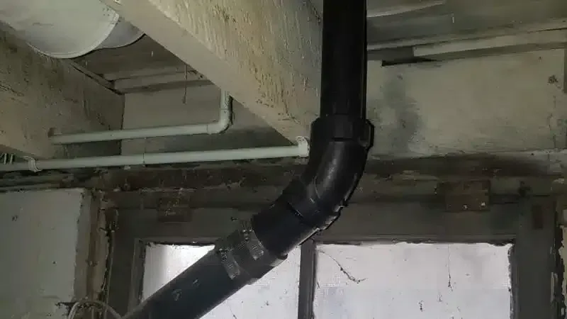Black plumbing pipes with a bend are installed on a concrete wall and ceiling, near a window.