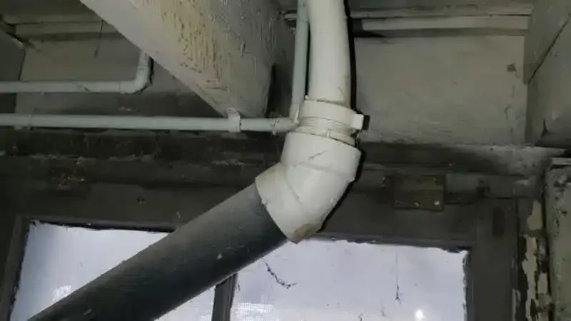 Pipes in a room, with a gray pipe descending, connected by white elbow joint and clamp. Window below.