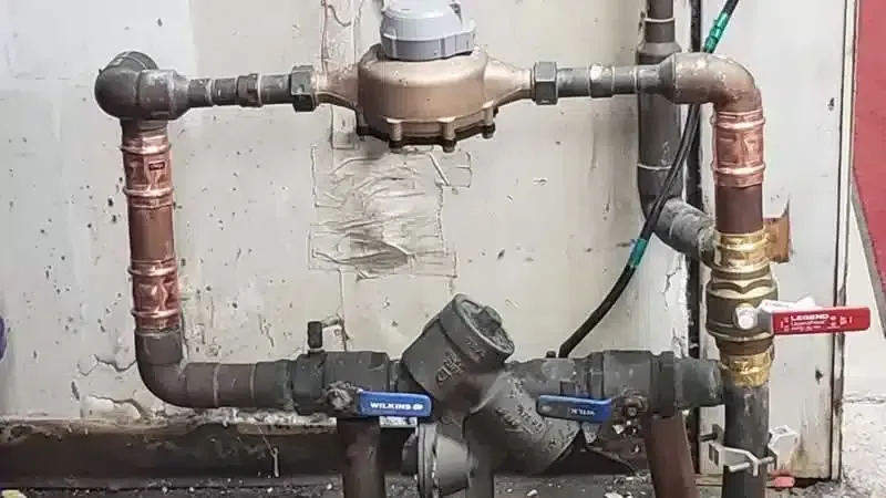 Copper and black plumbing pipes with a water meter and valves against a worn wall.