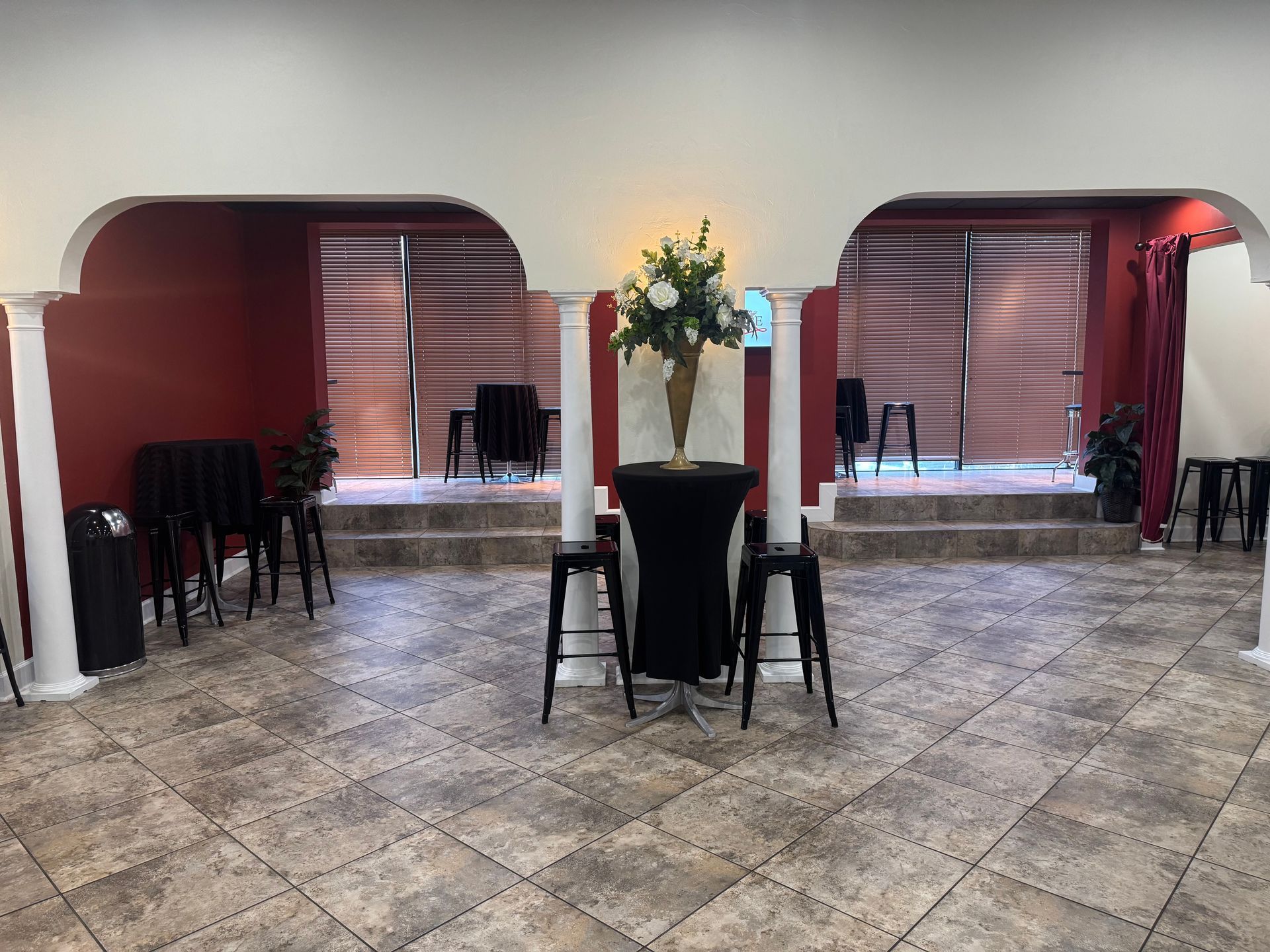 Indoor event hall with tiled floor, red drapes, and tall center table with flower arrangement and stools