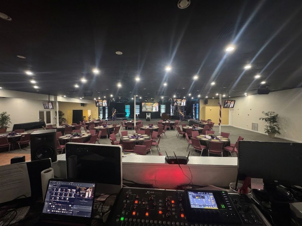 Empty banquet hall viewed from a DJ booth, with rows of tables and chairs under bright ceiling lights.