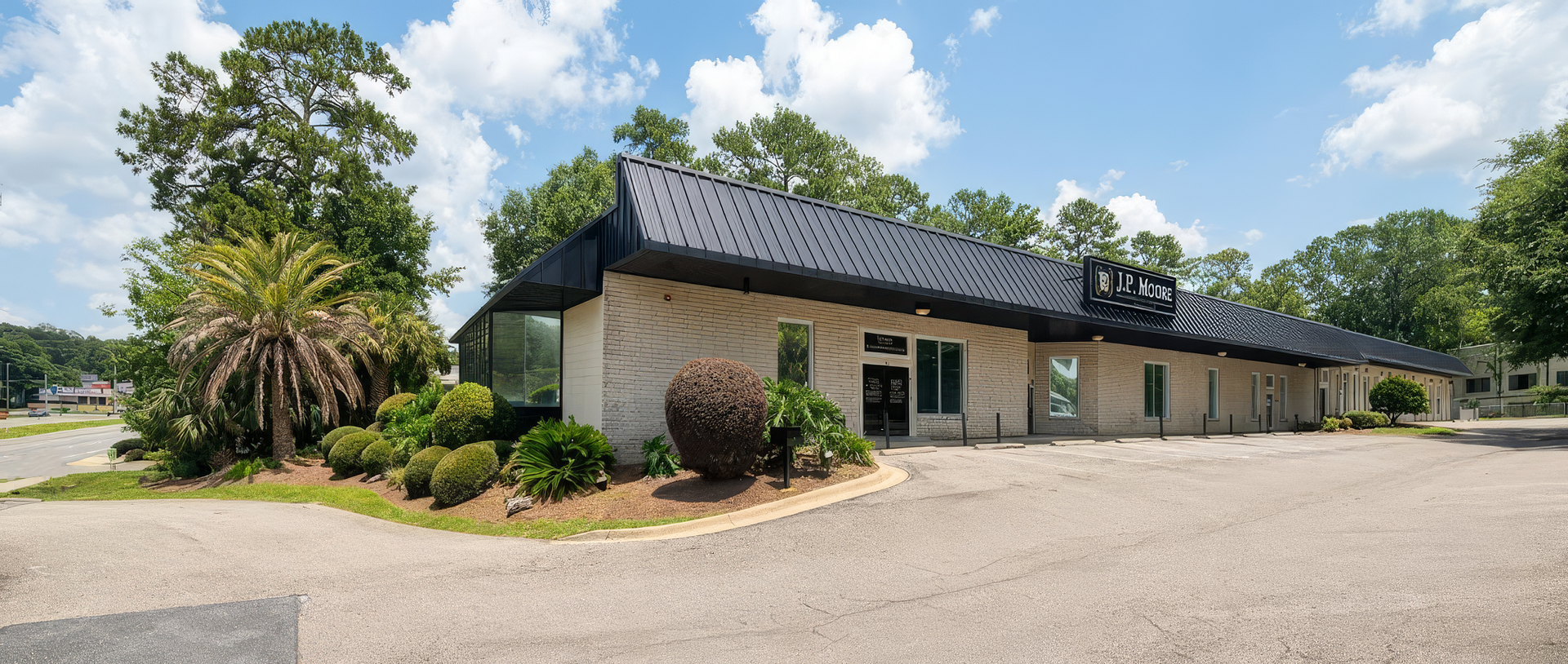 Single-story beige commercial building with dark roof, surrounded by trees and shrubs on a sunny day
