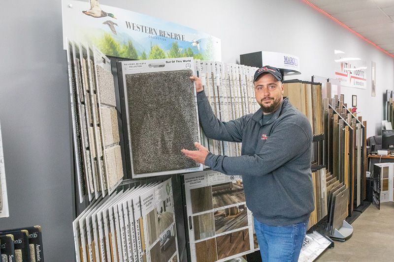 Man in a store holding a dark gray tile sample, pointing to it. Displays of tiles are behind him.