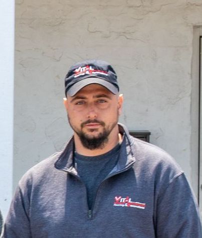 Man wearing a cap and gray shirt with a logo, standing outdoors.
