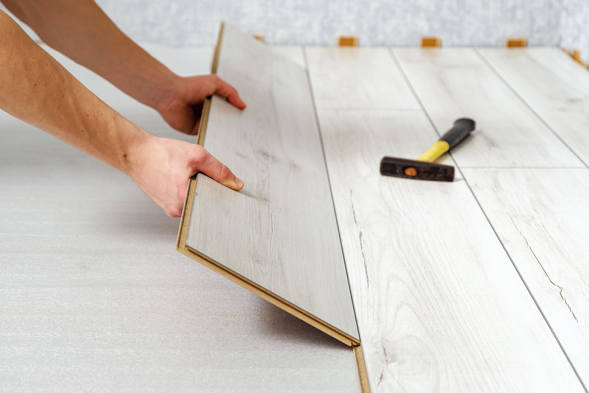 Hands installing a light-colored laminate floor, with a hammer nearby.