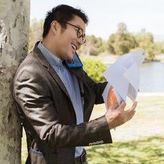 An image man leaning on a tree answering a phone while holding a bunch of papers