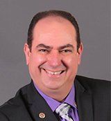 Man in suit smiling, purple shirt, patterned tie, against a gray background.