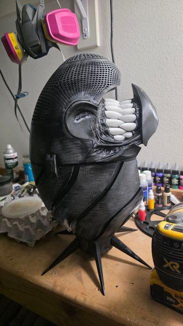 Black 3D-printed cosplay helmet with white teeth, hand-finished and displayed on a spiked stand