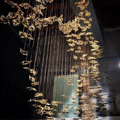 A large, dramatic chandelier composed of numerous golden, leaf-like shapes suspended by fine wires in a dark room.