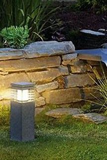 A glowing square-topped garden light stands in a grassy yard in front of a natural stone retaining wall at dusk.