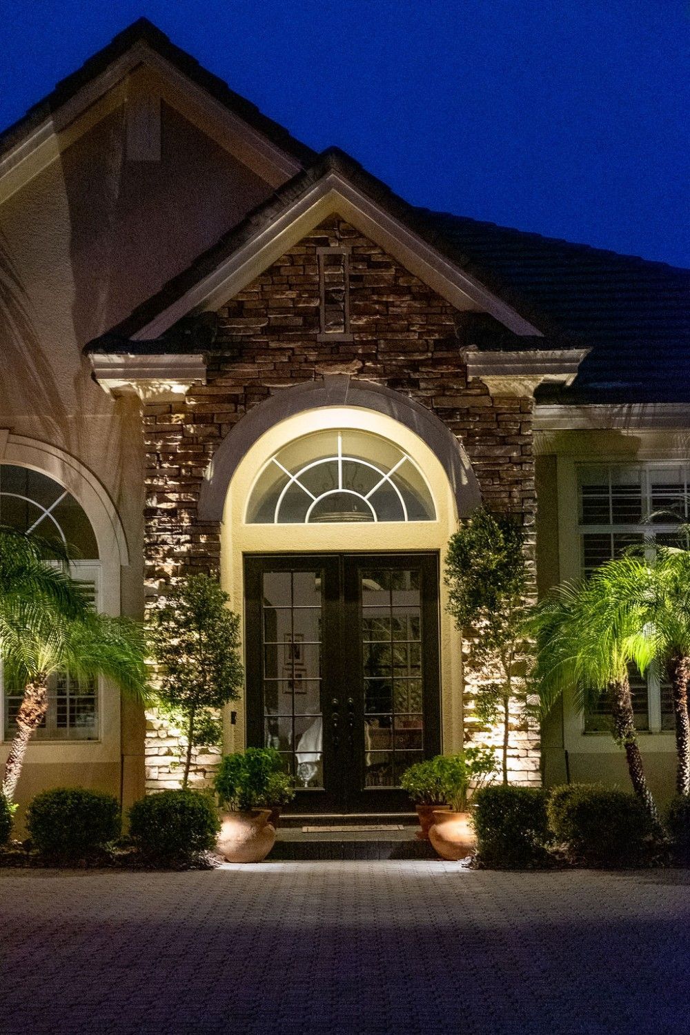 A lit stone entrance of a house at dusk with arched windows, double doors, and potted plants flanking the doorway.