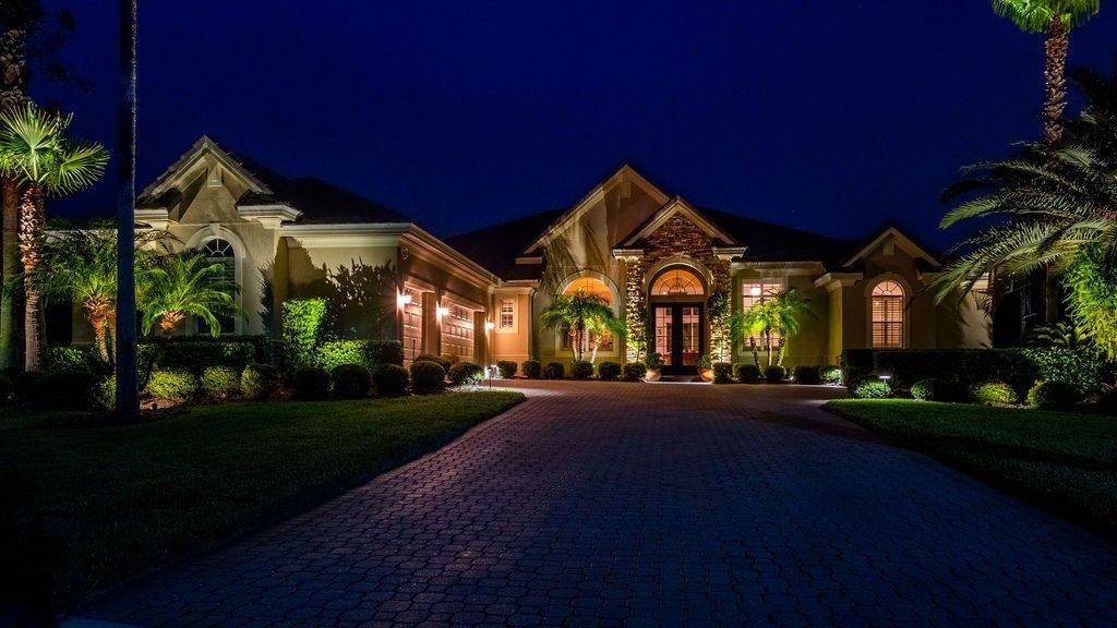 A luxurious single-story home at night with illuminated landscaping and a paved driveway under a dark blue sky.