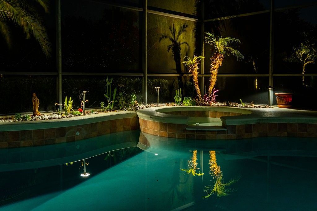 An illuminated backyard pool and hot tub at night, with palm trees and landscaping reflected in the calm water.