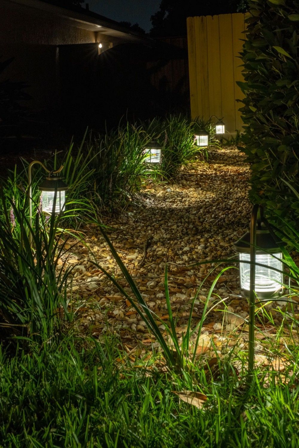 A line of small, glowing solar lanterns placed along a dark, leaf-covered garden path at night.