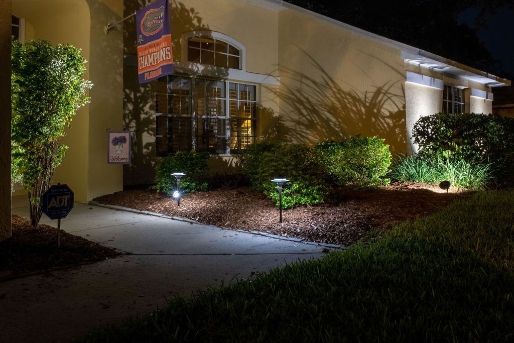 A yellow house exterior at night, illuminated by soft landscape lights along a walkway and against the bushes.