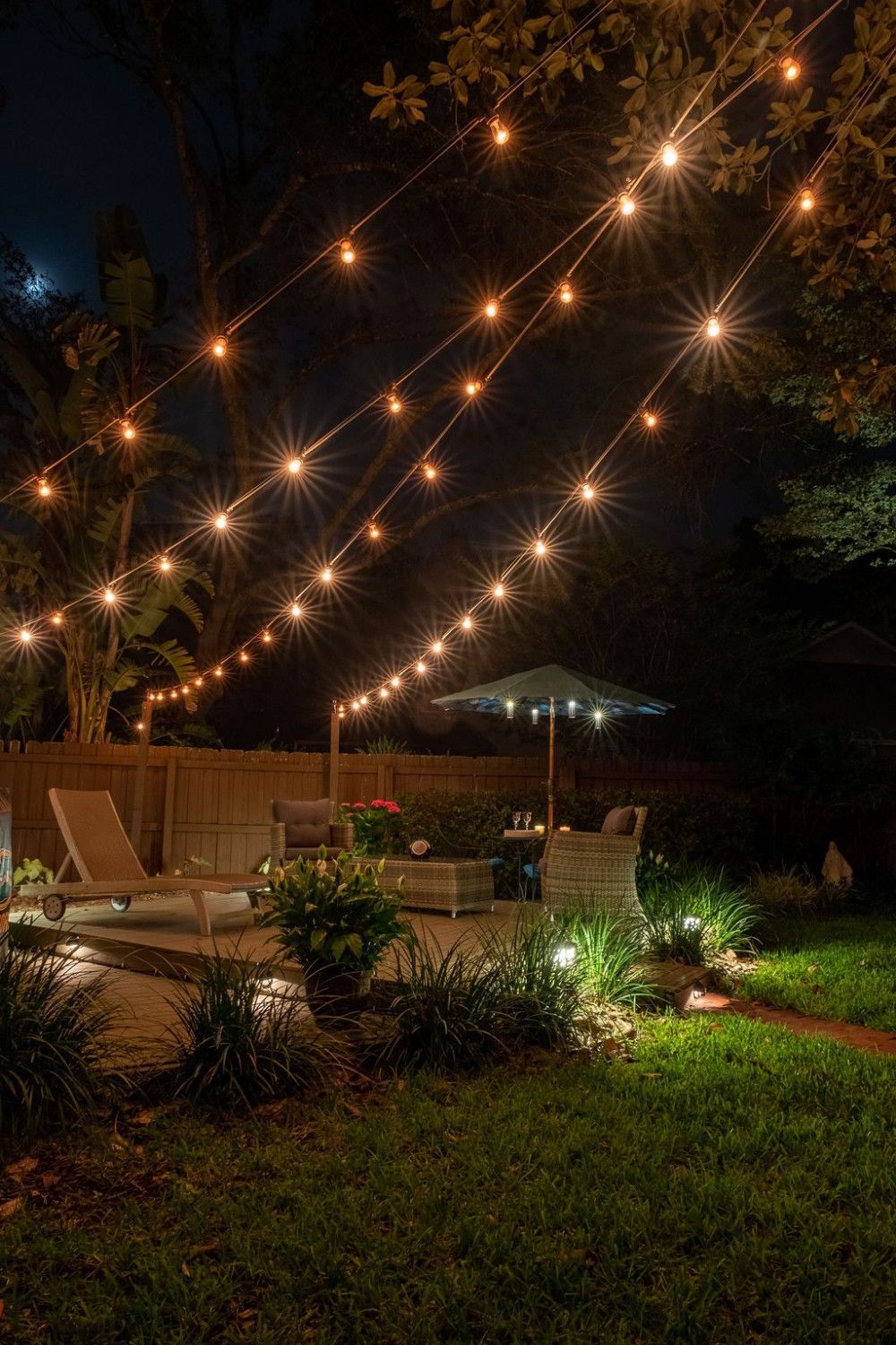 A cozy backyard patio at night, illuminated by warm string lights hung above lounge chairs and an umbrella.