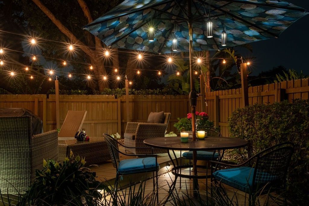 An outdoor patio at night featuring a table with an umbrella, chairs, and string lights hung along a wooden fence.