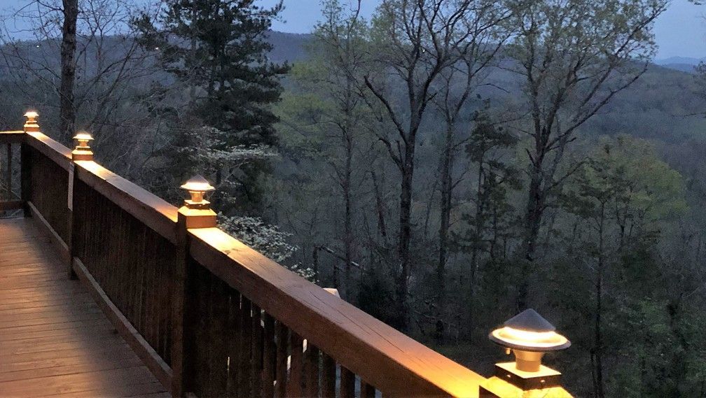 A wooden deck railing with glowing post cap lights overlooking a forest at dusk.