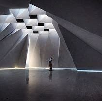 A person stands in a large, minimalist space with angular, geometric concrete walls and light filtering from the ceiling.