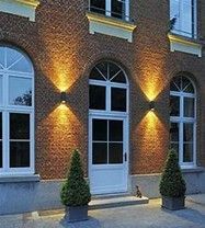 A brick building facade at night featuring a central white door, two flanking windows, and two glowing wall sconces.
