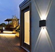 An exterior wall lamp shines a dual-beam, X-shaped light pattern onto a modern building entrance during dusk.