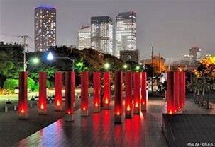 Rows of illuminated red pillars stand on a wooden deck at dusk, with tall city skyscrapers visible in the background.